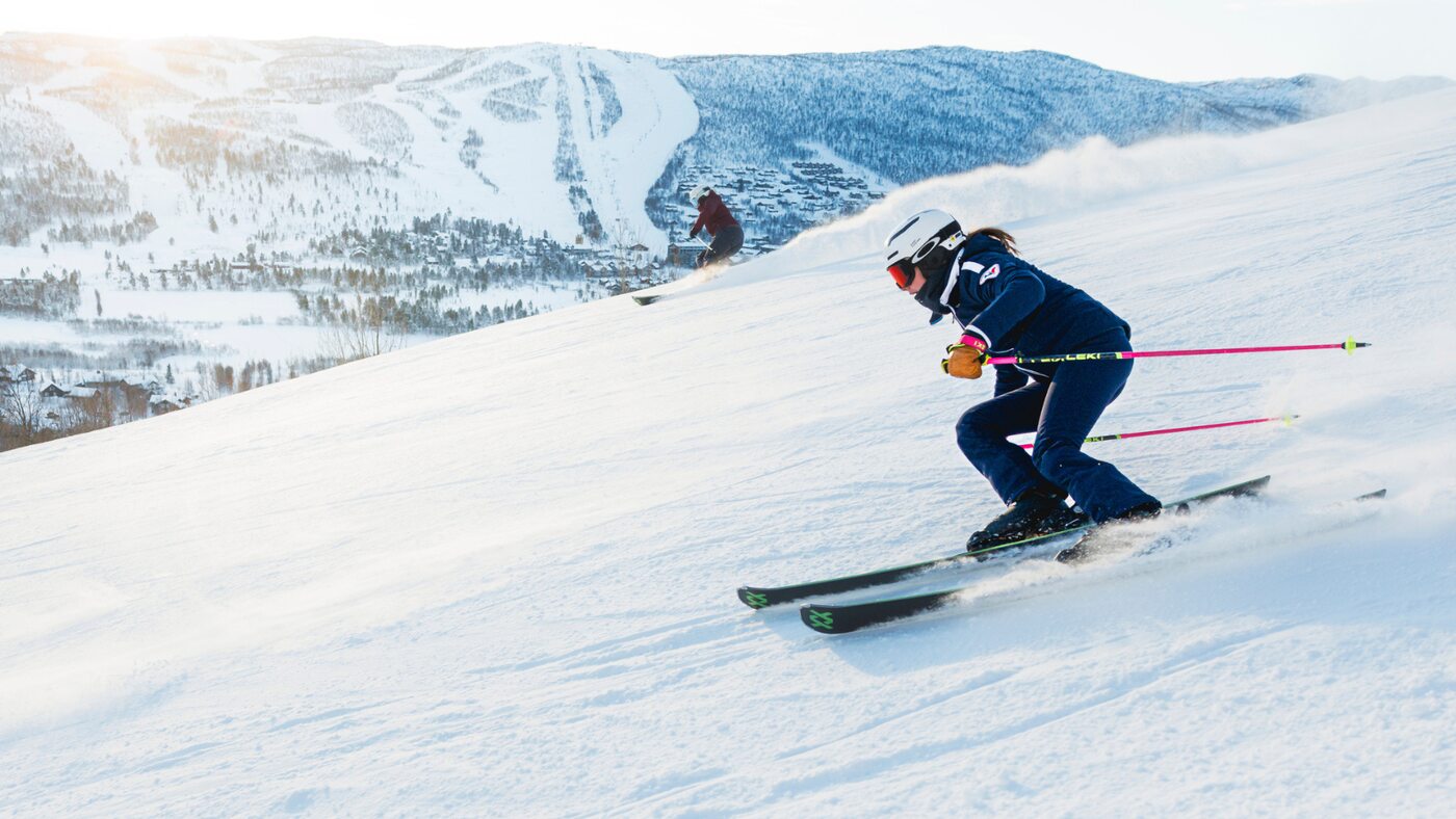 A skier descending a sunlit snowy slope above the valley and forested ski runs in Geilo