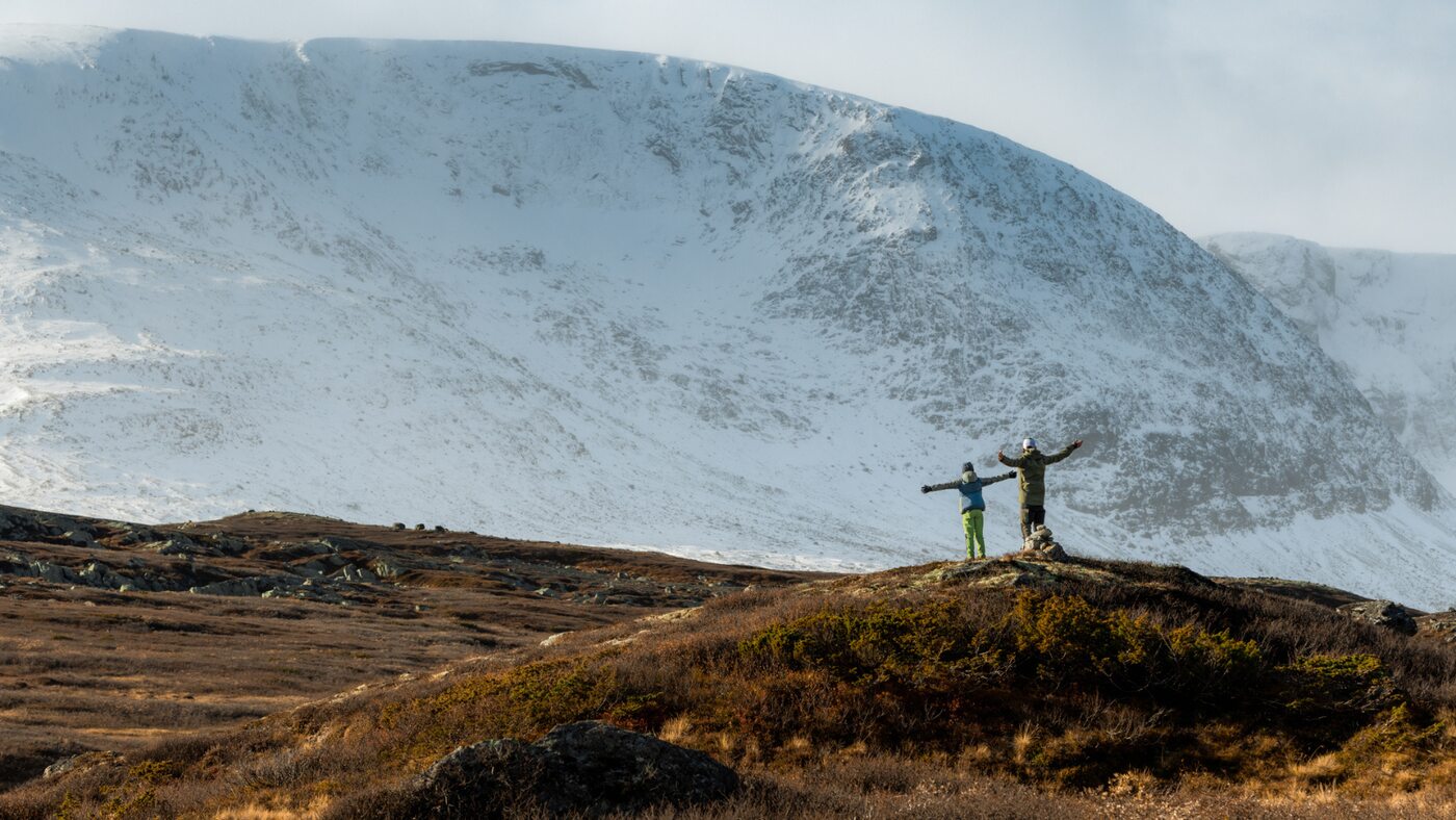 Two walkers on autumn-coloured ground below a snow-covered mountain ridge